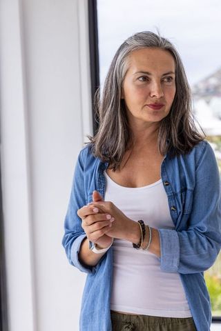 Contemplating middle-aged woman by window wearing denim shirt, smartwatch, casual home style
