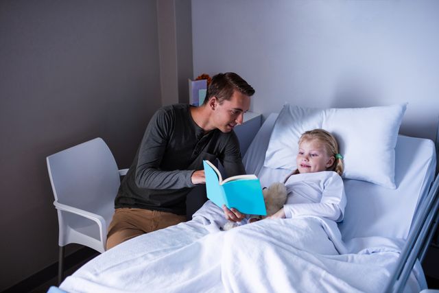 Happy girl on a bed reading book with her father in hospital