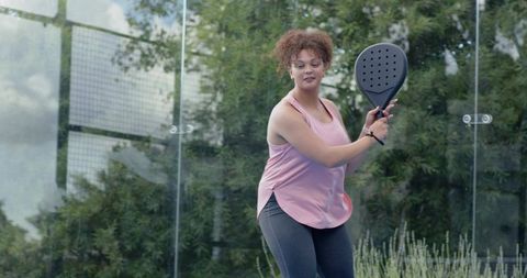 Female athlete playing padel on glass court wearing pink tank top and perforated racket