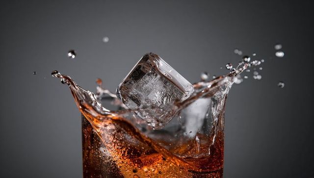 Ice cube splashing into cola glass creating crown splash and effervescent bubbles
