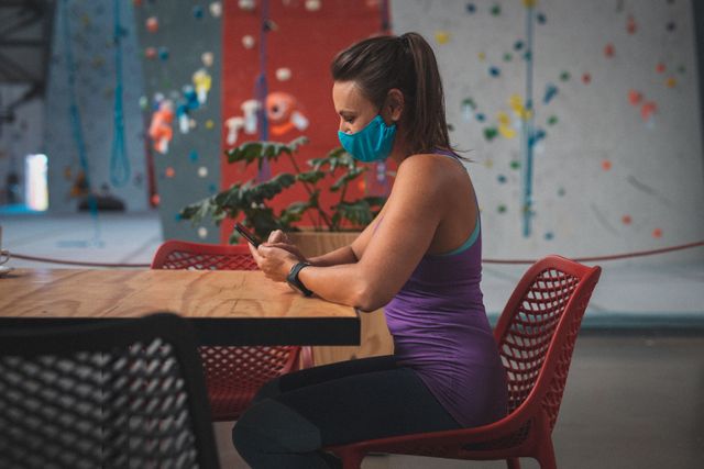 Caucasian woman wearing face mask sitting at table using smartphone in indoor climbing gym. Ideal for content related to fitness, health and safety during the COVID-19 pandemic, active lifestyle, and technology use in leisure activities.
