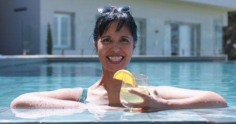 Smiling woman relaxing in pool holding lemon cocktail and sunglasses at sunny resort
