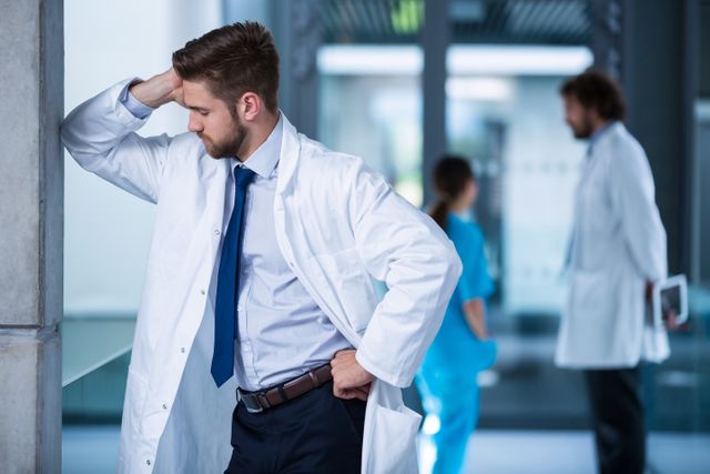 Doctor in white coat leaning against wall in hospital corridor, appearing stressed and anxious. Background includes other medical professionals, suggesting a busy healthcare environment. Useful for illustrating themes of medical stress, healthcare challenges, and the demanding nature of medical professions.