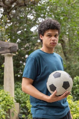 Young man standing in backyard holding soccer ball near stone column and lush greenery