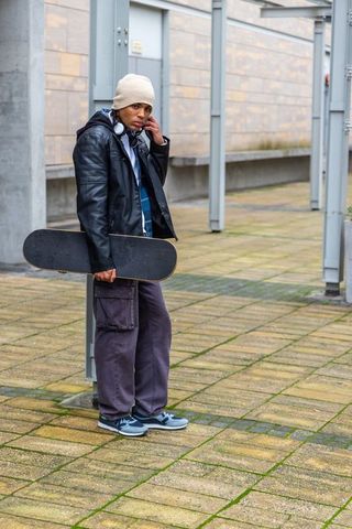 Urban skateboarder holding deck checking smartphone leaning on metal post in courtyard
