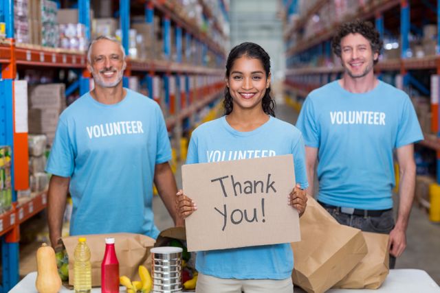 Group of volunteers standing in a warehouse, holding a thank you sign. They are smiling and wearing blue shirts with 'Volunteer' written on them. The warehouse shelves are stocked with various items, indicating a setting for community service or a food bank. This image can be used for promoting volunteer work, charity events, community service programs, and humanitarian aid campaigns.