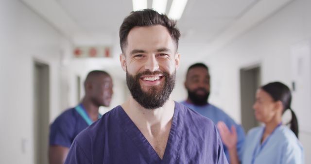 Male nurse wearing purple scrubs smiling confidently in a hospital corridor with diverse colleagues in the background engaging in discussion. Ideal for medical and healthcare promotional materials, teamwork and collaboration concepts, or nursing-focused content.