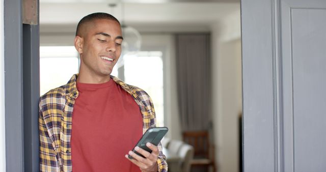 Young man leaning against door frame inside home, smiling while looking at smartphone. Ideal for promoting modern lifestyles, smart home features, or illustrating relaxed technology use at home.