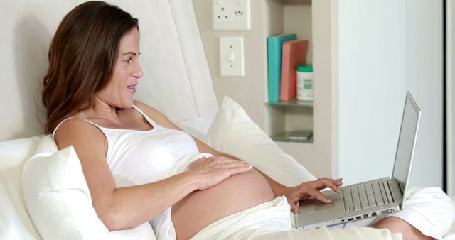 Smiling caucasian pregnant woman holding her belly using laptop at home, copy space. Pregnancy, motherhood, domestic life and wellbeing concept, unaltered.