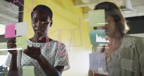 Female Colleagues Collaborating with Sticky Notes and Data Charts on Glass Wall