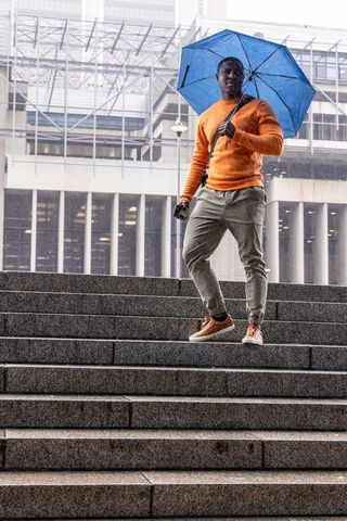 Urban commuter wearing orange sweater descending wet steps holding blue umbrella checking smartphone