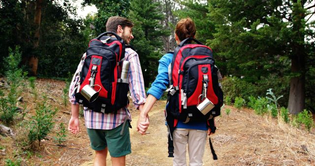 A young Caucasian couple is hiking in a forested area, with copy space. They are holding hands and carrying backpacks, suggesting an adventurous outdoor activity together.