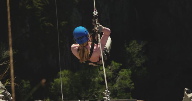 A woman with a blue helmet and safety harness riding a zip line over a lush forest. Ideal for promoting outdoor adventure activities, eco-tourism, safety equipment, and recreational sports. Shows the exhilarating experience of zip lining and emphasizes outdoor fun and nature exploration.