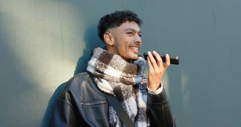 Young man leaning on teal wall speaking to phone wearing leather jacket and checked scarf