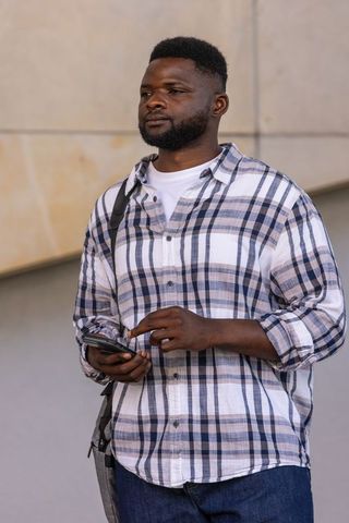 African man standing holding smartphone by beige wall in plaid shirt and crossbody bag