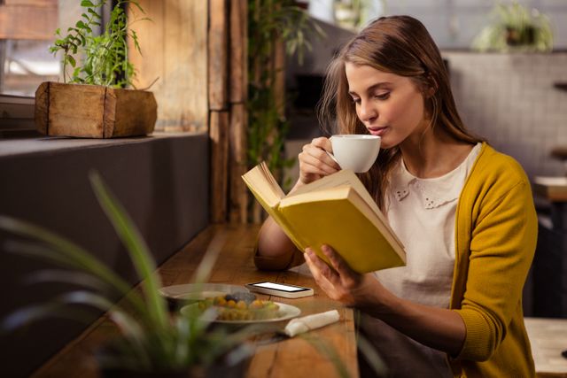 Woman enjoying a quiet moment reading a book and sipping coffee in a cozy cafe. Ideal for use in lifestyle blogs, coffee shop promotions, relaxation and leisure articles, and advertisements for cafes or bookstores.