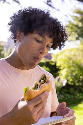 Young man enjoying sesame sandwich outdoors in sunlit backyard garden close-up summer lunch