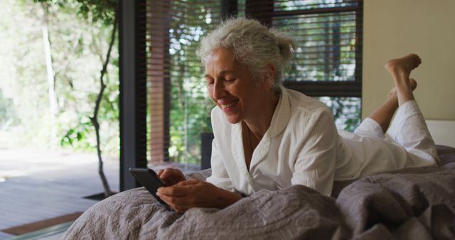 Mature woman happily using smartphone while relaxing on bed. Perfect for depicting technology usage among seniors, home relaxation, and joyful moments in everyday life.