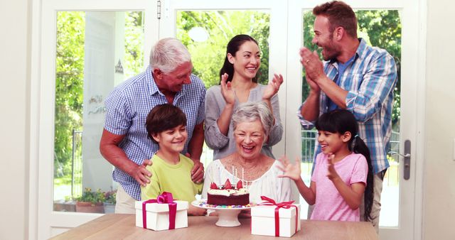 Multi-generational family gathered around a table celebrating grandmother's birthday. Children present wrapped gifts while grandmother smiles receiving a cake with candles. Suitable for use in advertisements, social media, blog posts, and articles about family events, celebrations, and unity.