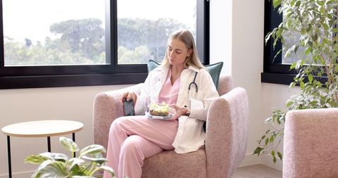 Female nurse in pink scrubs taking lunch break in pastel clinic lounge holding salad and smartphone