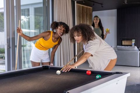Multicultural women playing pool at home on modern black billiard table enjoying competition