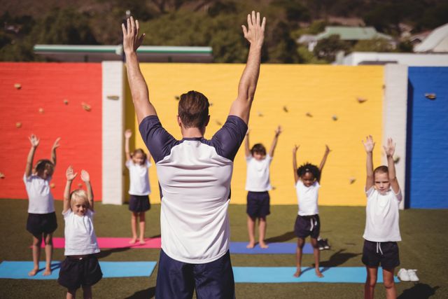 Coach leading a group of school kids in an outdoor exercise session. Children are following the coach's instructions, stretching their arms upwards. Ideal for use in educational materials, fitness programs, health and wellness campaigns, and advertisements promoting physical activity for children.