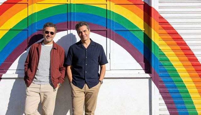 Two men leaning against rainbow mural on sunny urban wall, casual lifestyle portrait