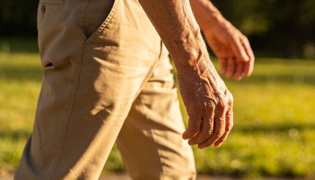 Walking senior man showing wrinkled hands wearing khaki trousers in golden hour park