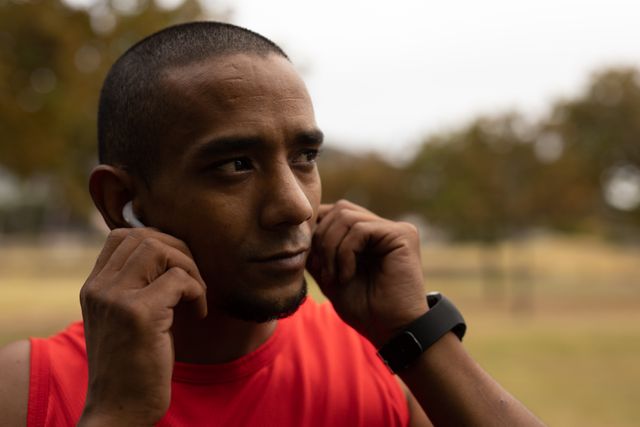 Biracial man in red sportswear and smartwatch adjusting wireless earphones while taking a break from running workout in a park. Ideal for use in fitness, technology, and healthy lifestyle promotions, as well as advertisements for sportswear, wearable tech, and outdoor activities.