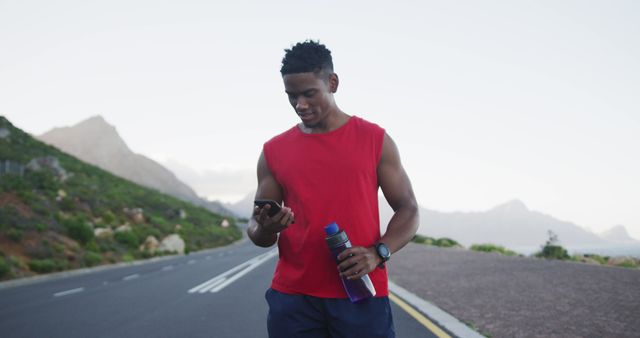 African american man holding water bottle using smartphone on the road. fitness sports and healthy lifestyle concept