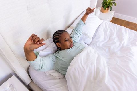 African american woman stretching in sunlit bedroom with white bedding and bedside plant