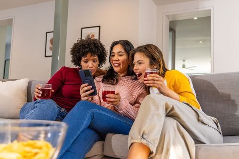Diverse women on sofa watching smartphone together while holding drinks and sharing snacks