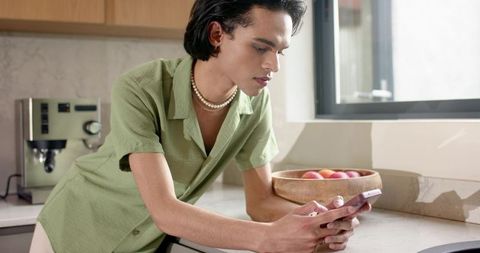 Young man checking smartphone by bright marble kitchen counter with espresso machine and fruit bowl