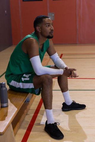 African american basketball player sitting on bench wearing green jersey resting on court