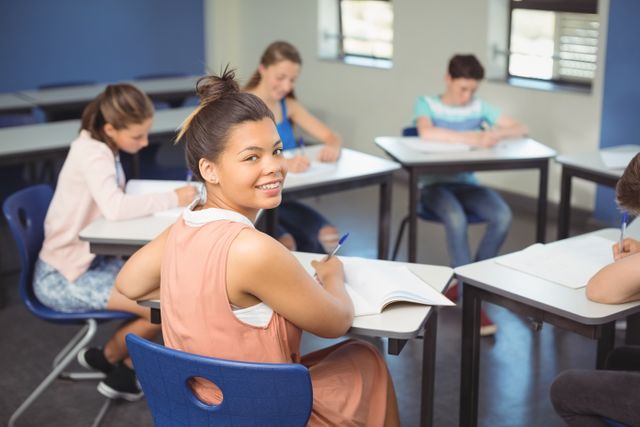 Schoolgirl smiling while sitting at desk in classroom with other students. Ideal for educational materials, school brochures, and websites promoting academic programs. Highlights positive learning environment and student engagement.