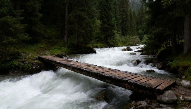 Rustic wooden plank footbridge crossing rushing alpine river through mossy conifer forest