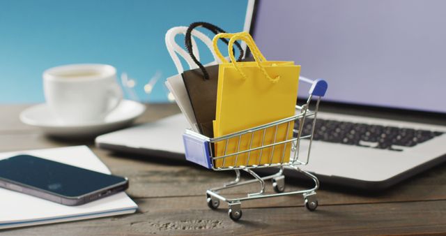 Mini shopping cart filled with colorful shopping bags sits on a wooden desk next to laptop, smartphone, coffee cup. Great for illustrating concepts like online shopping, e-commerce platforms, digital retail, consumer habits, and technology in business. Ideal for use in blog articles, e-commerce websites, digital marketing materials, and online business promotions.