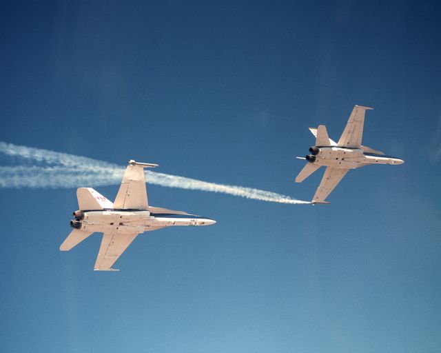 Two NASA F/A-18 jets are captured during an Autonomous Formation Flight (AFF) program experiment. Visible smoke generators outline the twisting paths of wingtip vortices behind the aircraft. This image can be used in aerospace research discussions, aviation technology articles, and educational materials about flight dynamics and experimental aircraft.