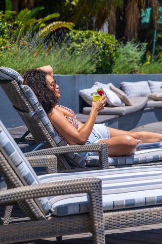 African american woman relaxing poolside on lounger sipping cocktail in tropical sunshine