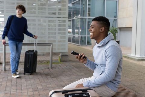 Diverse male travelers chatting and smiling at urban plaza with suitcases and smartphone