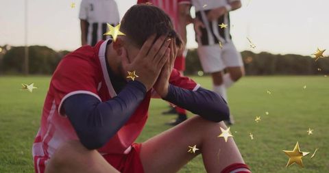 Sitting soccer player covering face on grass pitch wearing red kit and long-sleeve base