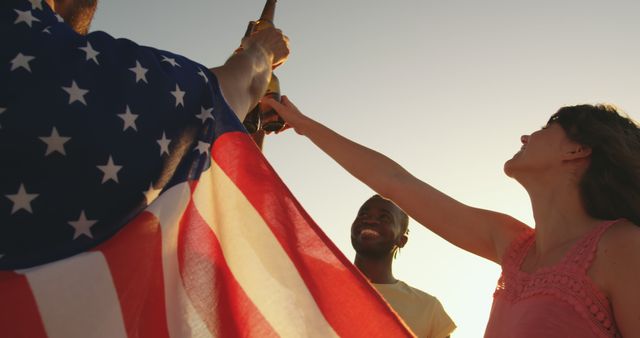 This picture shows friends toasting with beers at sunset while one wears a USA flag. Great for depicting unity, diversity, and celebration during outdoor summertime gatherings. Perfect for marketing materials related to parties, holidays, or American culture events.