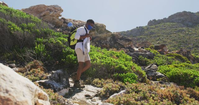 An adventurous young man is trekking on a rocky mountain trail surrounded by lush greenery. Ideal for promoting outdoor activities, nature trips, fitness, and exploration. Perfect for blog posts, travel magazines, adventure guides, and advertisements targeting outdoor enthusiasts and adventure-seekers.