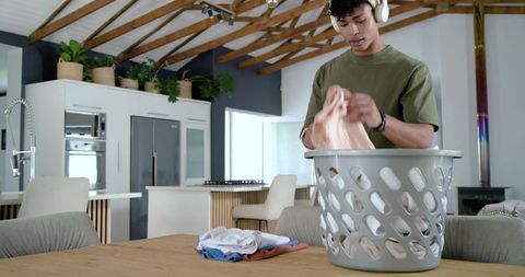 Young man folding laundry in modern open-plan kitchen wearing headphones, sunlit interior