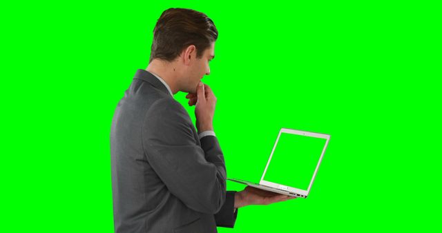 Businessman wearing a suit, holding a laptop with a green screen background, deep in thought. Useful for technology, business, and corporate marketing materials. Ideal for creating digital content, presentations, and advertisements depicting professional environments.