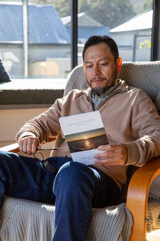 Mature asian man reading sunset booklet in sunlit armchair, holding glasses, cozy home