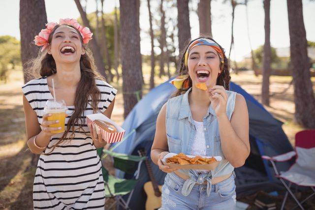 Two young women are enjoying snacks and drinks while camping in a forest on a sunny day. They are smiling and appear to be having a great time. This image can be used for promoting outdoor activities, camping gear, summer vacations, and lifestyle blogs focused on adventure and leisure.