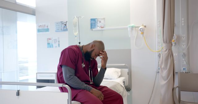 Medical professional wearing scrubs holding head while sitting on hospital bed, indicating stress and fatigue. Hospital equipment and patient bed in background emphasizing healthcare setting. Suitable for themes of medical burnout, healthcare stress, mental health in healthcare, overworked nurses or doctors, hospital environment, and the emotional toll on healthcare workers.