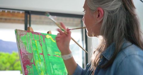 Mature woman painting vibrant green and pink abstract canvas at easel in sunlit studio