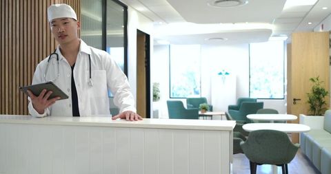 Asian doctor standing behind clinic reception desk holding tablet in modern bright lobby